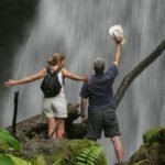 This image depicts a confident woman hiking through a verdant rainforest toward a cascading waterfall, embodying the essence of sustainable travel. She carries a lightweight backpack filled with eco-friendly gear, dressed in ethical activewear, surrounded by pristine natural scenery that highlights biodiversity conservation. The scene captures the empowerment of solo exploration in harmony with the environment, with soft sunlight filtering through the canopy to evoke tranquility and responsibility. Sustainable Travel Products Revolutionizing Women's Journeys