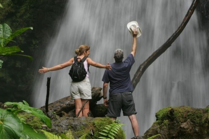 sustainable-women-travel-hero This image depicts a confident woman hiking through a verdant rainforest toward a cascading waterfall, embodying the essence of sustainable travel. She carries a lightweight backpack filled with eco-friendly gear, dressed in ethical activewear, surrounded by pristine natural scenery that highlights biodiversity conservation. The scene captures the empowerment of solo exploration in harmony with the environment, with soft sunlight filtering through the canopy to evoke tranquility and responsibility. Sustainable Travel Products Revolutionizing Women's Journeys